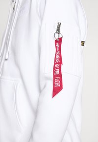 Veste à capuche blanche zippée avec une poche dotée d'une fermeture éclair argentée et d'une étiquette en ruban rouge portant l'inscription "ENLEVEZ AVANT LE VOL". Texture uniforme, design décontracté.