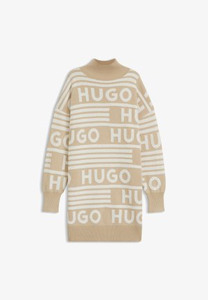 Beige gebreide trui met wit "HUGO" logo patroon, hoge hals, en een losse pasvorm, met ribboorden aan de mouwen en onderkant.