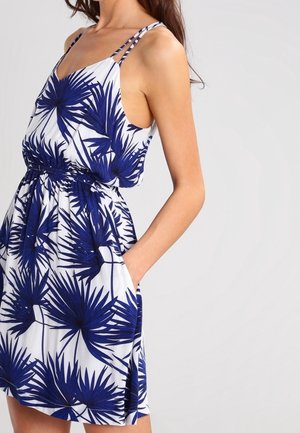 Frau trägt ein weißes ärmelloses Kleid mit blauem Palmenblattmuster und elastischem Taillenbund, Hand in der Tasche, vor einfarbigem Hintergrund.