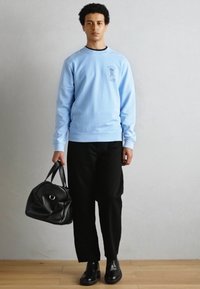 Light blue sweatshirt with a circular logo, black pants, and shiny black shoes. Holding a black duffel bag, standing on a wooden floor.