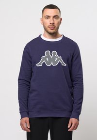 Námornícky modrý sweatshirt vyrobený z bavlny s okrúhlym výstrihom. Na prednej strane sa nachádza veľké šedé a biele grafické logo. Dlhé rukávy.