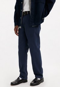 Pantalon bleu foncé en tissu tissé avec une coupe droite, associé à des mocassins noirs et une veste en denim portée sur un t-shirt gris.