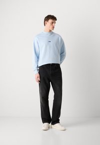 Felpa azzurra con scollo rotondo e piccolo logo ricamato, abbinata a jeans neri e sneaker crema. In piedi su uno sfondo semplice.