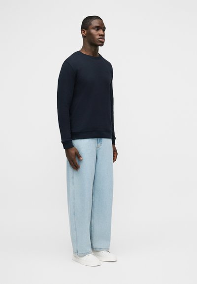 Άνδρας στέκεται προς τα αριστερά φορώντας μπλε σκούρο φούτερ, ανοιχτό μπλε τζιν με φαρδύ πόδι και λευκά αθλητικά παπούτσια, σε απλό γκρι φόντο.