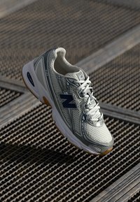 Zapatilla de running New Balance en blanco y gris con logo azul sobre un fondo de rejilla de metal texturizada.