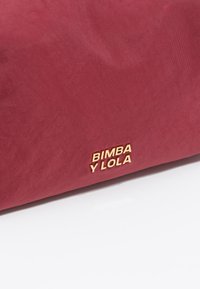 Bolso de tela roja con una textura suave y un logo dorado en relieve "BIMBA Y LOLA" en un lado. Forma rectangular simple.
