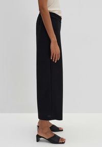 Pantalons noirs taille haute à jambes larges avec une texture lisse. Dotés d'une taille élastique et de poches latérales. Associés à des sandales noires à talons.