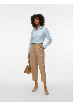 Femme aux cheveux bouclés portant une chemise bleu clair, un pantalon beige ceinturé, tenant un sac à main marron et portant des sandales à talons claires.