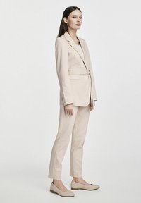 Beżowy dopasowany garnitur z marynarką na pasek i dopasowanymi spodniami, zestawiony z pasującymi butami na niskim obcasie. Gładki materiał i minimalistyczny design.