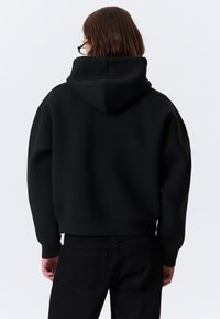Felpa nera con cappuccio, dal taglio comodo, con polsini a costine e tasca a marsupio. Indossata con pantaloni neri, dal texture liscia.