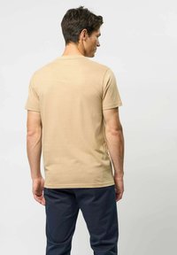 Egy férfi áll, háttal a kamerának, sima bézs rövidujjú pólóban és sötét navy színű nadrágban, egy világos háttér előtt.