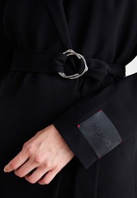 Sort frakke med et sølvfarvet oval spænde og bælte, med en tekstureret sort lap med rød syning mærket "HUGO" på ærmet. Glat stof.