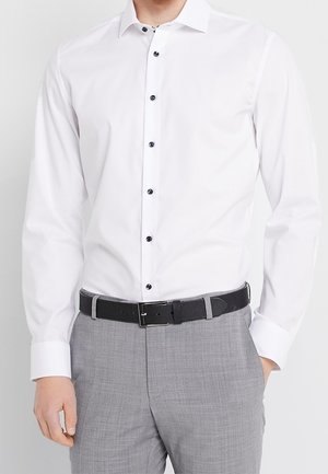 Mand iført en hvid langærmet skjorte med sorte knapper, grå bukser og sort bælte, står med den ene hånd i lommen.