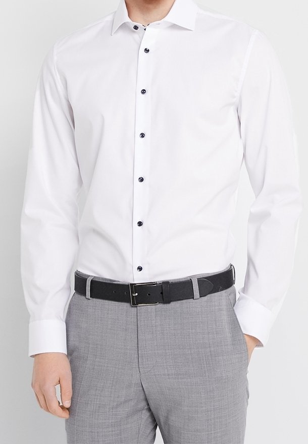 Homme portant une chemise blanche à manches longues avec des boutons noirs, un pantalon gris et une ceinture noire, debout avec une main dans la poche.