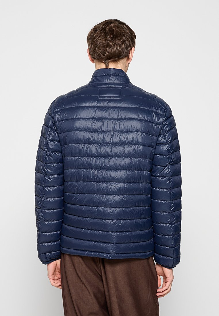 Hombre que lleva una chaqueta acolchada azul marino y pantalones marrones, visto desde atrás sobre un fondo liso.