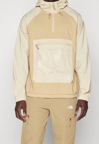 Suéter ligero beige con capucha, que presenta un bolsillo de malla, cierre de cremallera y puños elásticos. Pantalones beige a juego con cremalleras laterales.