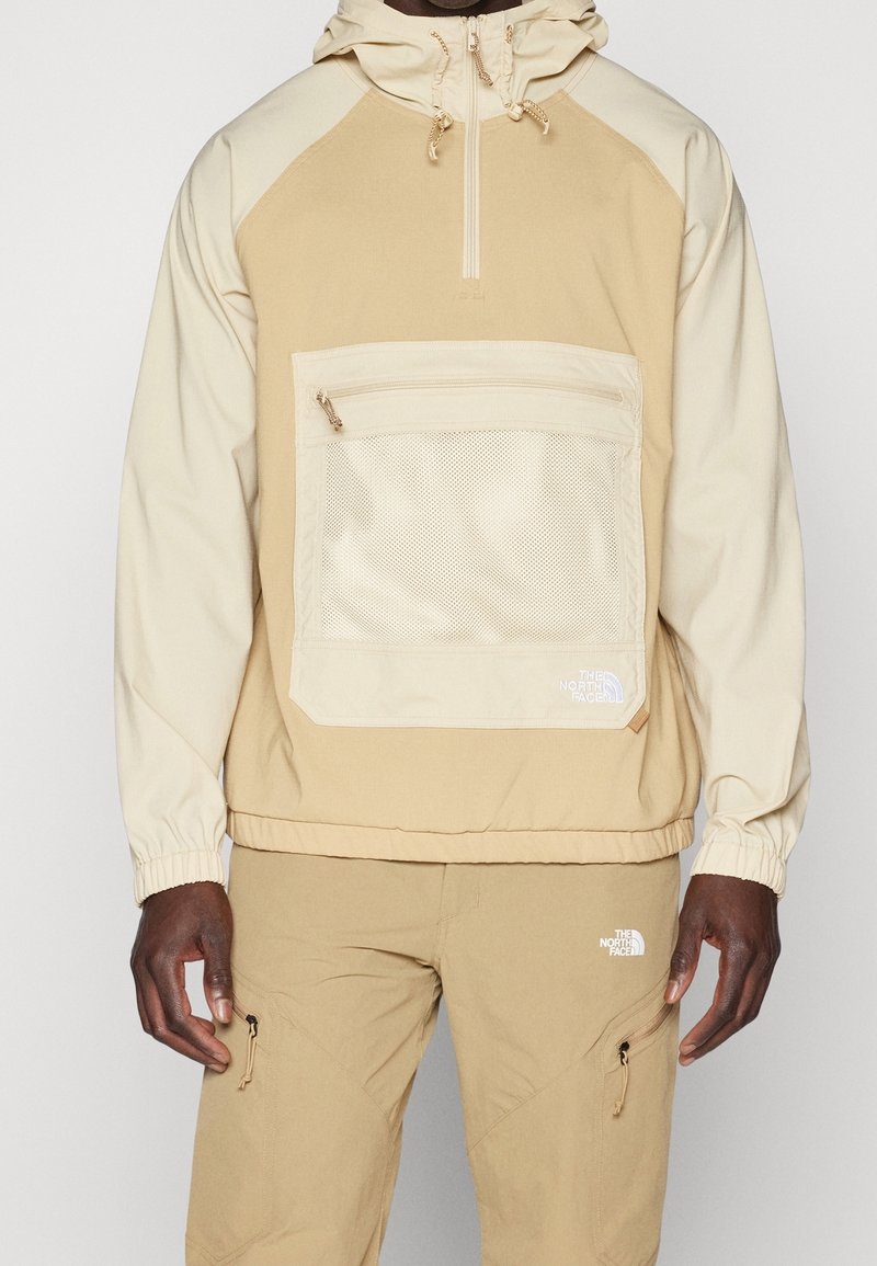 Suéter ligero beige con capucha, que presenta un bolsillo de malla, cierre de cremallera y puños elásticos. Pantalones beige a juego con cremalleras laterales.