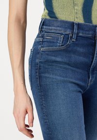 Skinny jeans made of dark blue denim, featuring a mid-rise waist, five pockets, and a small logo accent on the back pocket.