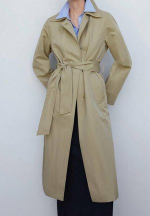 Osoba ubrana w beżowy płaszcz wiązany w pasie, na niebieskiej koszuli z kołnierzykiem i ciemnych spodniach, stojąca na jednolitym jasnym tle.