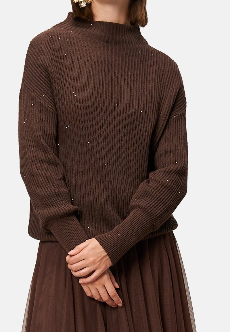 Femme portant un pull marron côtelé avec de petits sequins et une jupe plissée assortie, les mains doucement jointes devant.