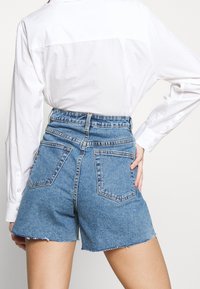 Shorts de mezclilla azul de talle alto con dobladillo deshilachado, que presentan un diseño clásico de cinco bolsillos y bolsillos traseros cosidos, combinados con una camisa blanca con cuello.