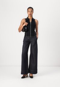 Schwarzes ärmelloses Zip-Top mit weißen Nähten, kombiniert mit dunklen, weiten Jeanshosen und schwarzen High Heels, während sie eine kleine schwarze Tasche hält.
