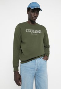 Olivovo zelená mikina s logom "GUESS EST. 1981", s rebrovanými manžetami a golierom. V kombinácii s svetlými modrými džínsami a svetlomodrou šiltovkou.