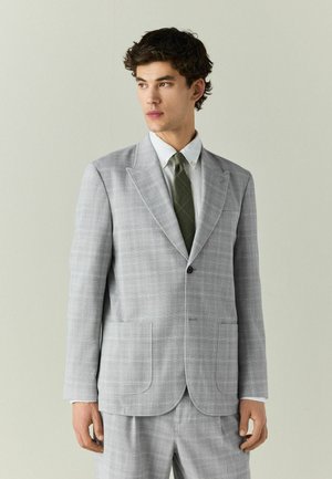 Junger Mann trägt einen hellgrauen karierten Anzug mit weißem Hemd und dunkelgrüner Krawatte, blickt seitlich vor einem schlichten hellen Hintergrund.