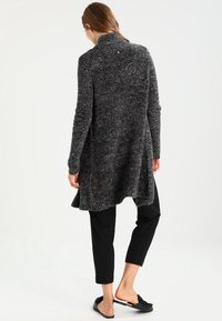 Langer, vorne offener Cardigan aus strukturiertem schwarzem und grauem Stoff, verlängert am Rücken, mit hohem Kragen und anliegenden Ärmeln, kombiniert mit schwarzen Hosen.