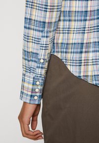 Flerfärgad rutskjorta med blå, gula och vita mönster, knäppning vid ärmen och rundad nederkant, matchad med bruna tygbyxor.