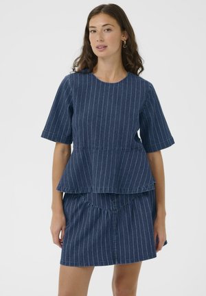 Mladá žena s vlnitými hnedými vlasmi, oblečená v modrom krátkym rukávom top s tenkými pruhmi a zodpovedajúcej sukni, stojaca na jednoduchom pozadí.