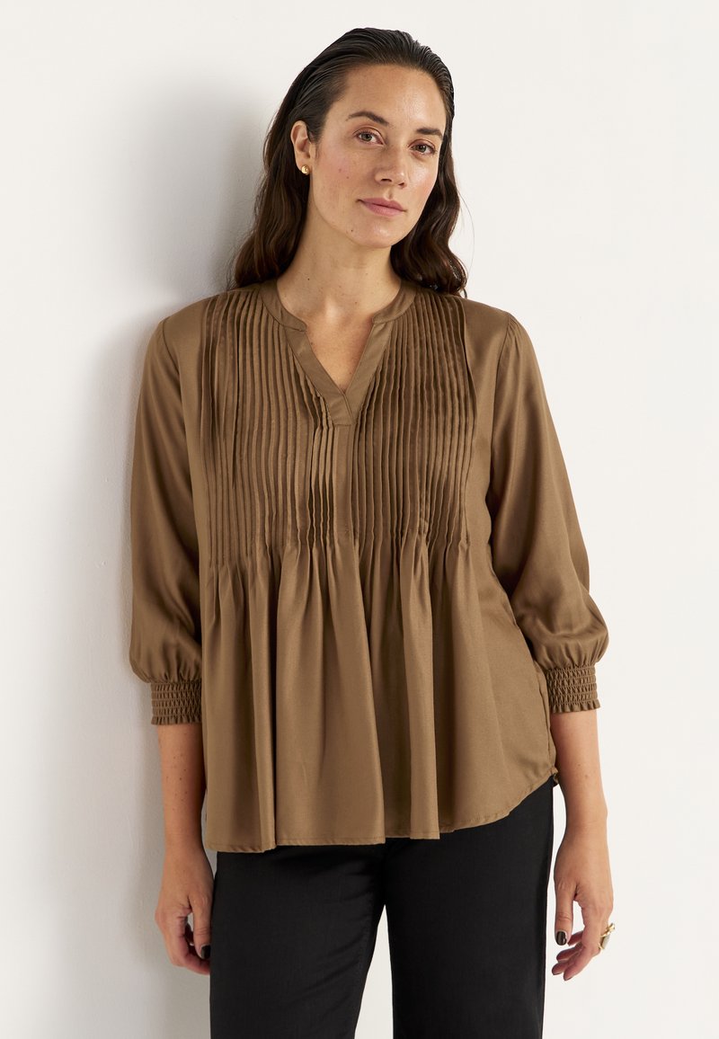Brun, langærmet bluse med V-hals og plisseret design. Har samlet detalje ved brystet og elastiske manchetter. Glat tekstur.