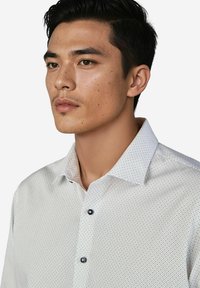 Junger Mann mit kurzen schwarzen Haaren und leichtem Gesichtshaar, der ein weißes Hemd mit kleinen schwarzen Punkten trägt und leicht nach links schaut.