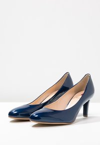 Marinblå lackläder högklackade skor med spetsig tå och 7,5 cm stilettklack. Inuti finns en ljusbeige innersula. Slät och glänsande textur.