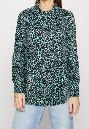 Mujer con camisa de manga larga abotonada con estampado de leopardo turquesa y negro y vaqueros azul claro.