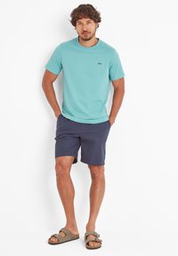 Man wearing a light blue short-sleeve t-shirt, navy shorts, and beige sandals standing with hands in pockets against white background.