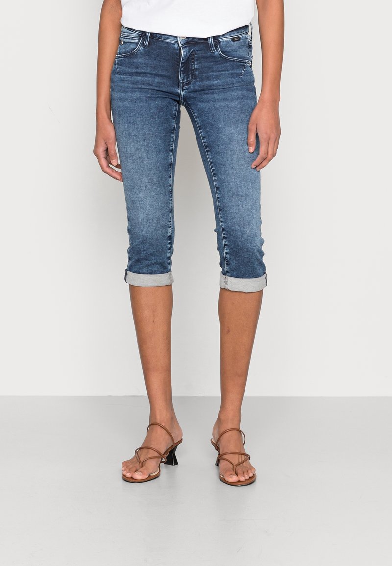 Femme portant un pantacourt en denim bleu retroussé et des sandales marron à talons avec lanières, debout devant un fond blanc.