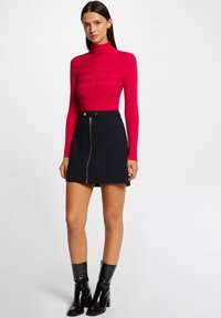 Top de manga larga rojo acanalado con cuello alto, combinado con una falda mini negra que tiene una cremallera frontal y bolsillos. Botines negros.
