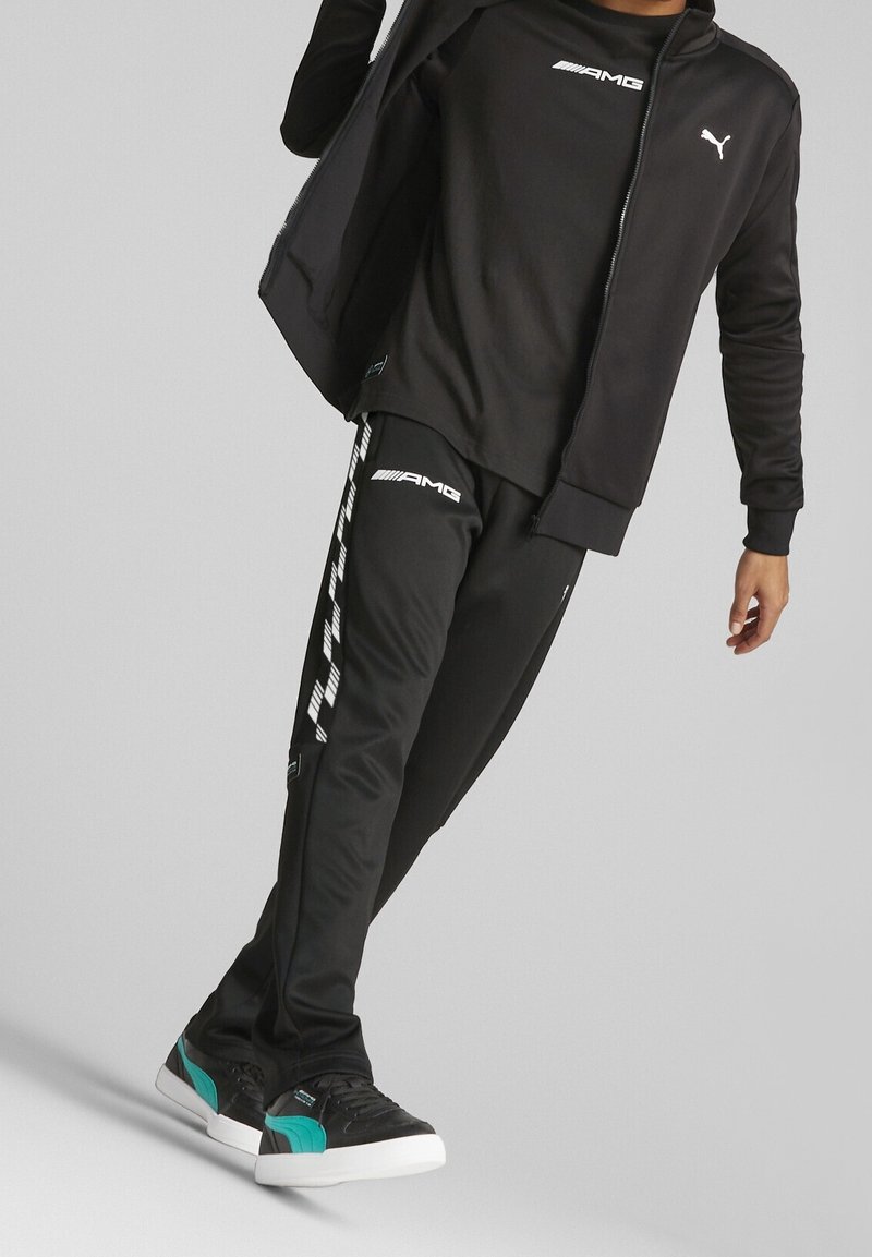 Personne portant une veste Puma noire, un t-shirt AMG noir, un pantalon de survêtement AMG noir avec des bandes blanches, et des baskets noires avec des accents bleu sarcelle.