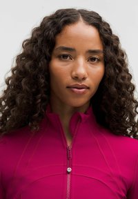 Kohandatud, kõrge kaelusega roosa tõmblukuga jakk, millel on kumerad õmblused ja väike logoaktsent tõmbel. Sile tekstuur ja kerge materjal.