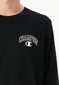 Juoda ilgo rankovės marškinėliai su balta "CHAMPION" užrašu ir logotipu viršutinėje kairėje krūtinės pusėje, dėvimi asmens priešais baltą foną.