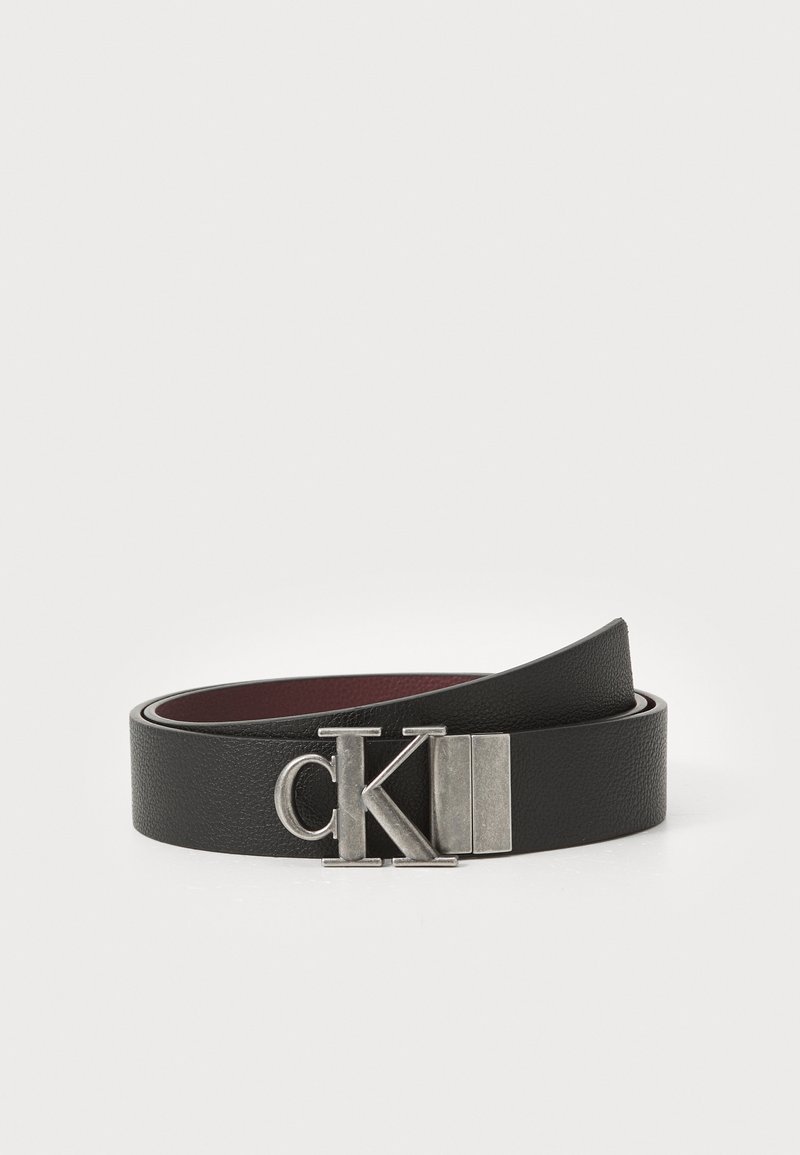 Zwarte leren riem met een gestructureerde afwerking, voorzien van een zilveren metalen gesp met de letters "CK". Van binnen heeft hij een donkerrood kleur.