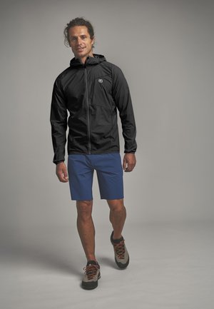 Mann, der nach vorne geht, trägt eine schwarze Kapuzenjacke, blaue Shorts und beige Wanderschuhe mit orangefarbenen Schnürsenkeln vor einem schlichten grauen Hintergrund.