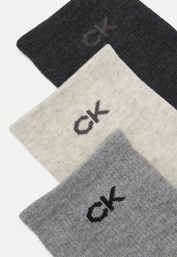 Tris kojinių porų: juodų, šviesiai pilkų ir tamsiai pilkų. Kiekvienoje poroje įsiuvinėta "ck" logotipo, išsiskiriančio kontrastingomis spalvomis. Minkšta, mezginio tekstūra.