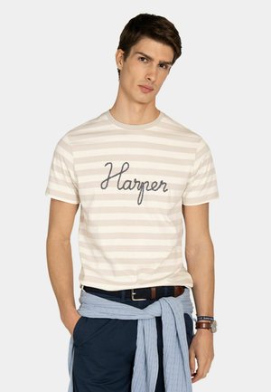 Joven con pelo corto castaño que lleva una camiseta beige con rayas y la palabra "Harper", pantalones azul marino y un suéter azul claro atado alrededor de la cintura.