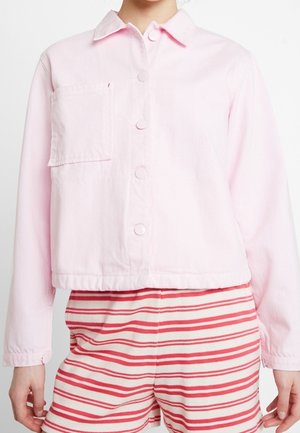 Osoba nosząca jasnoróżową kurtkę zapinaną na guziki z kieszenią na piersi oraz czerwono-białe, poziomo paskowane krótkie spodenki.
