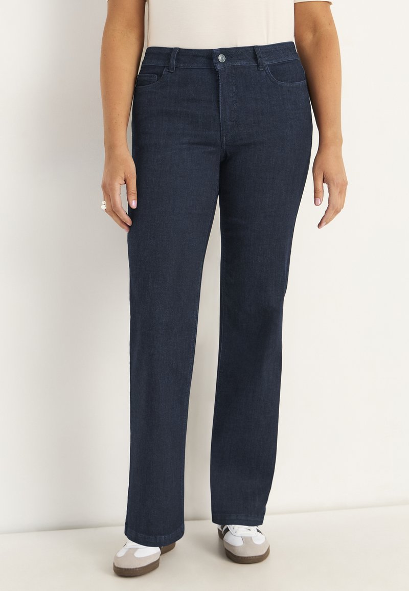Kvinna med mörkblå raka jeans och vita sneakers, ståendes mot en enfärgad ljus bakgrund.