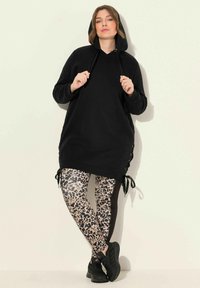 Černá kapucová tunika s bočními šňůrkami a dlouhými rukávy, kombinovaná s legínami s leopardím potiskem. Černé sportovní boty dotvářejí celý outfit.