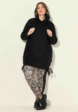 Schwarze Kapuzen-Tunika mit seitlichen Bändern und langen Ärmeln, kombiniert mit Leggings im Leopardenmuster. Schwarze Sportschuhe runden das Outfit ab.