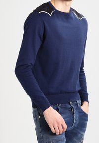 Pullover blu navy con maniche lunghe, polsi a coste e scollo rotondo; presenta accenti neri sulle spalle e dettagli di cucitura bianca.